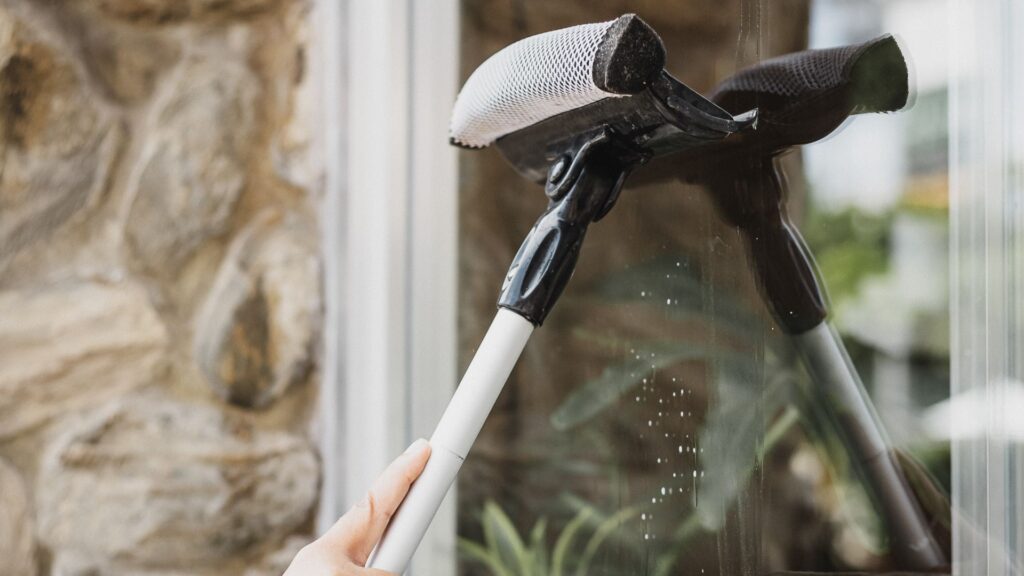 Persona utilizando un limpiador de ventanas con mango blanco y cabezal negro, limpiando un cristal con un paño blanco, reflejando el entorno con vegetación y una pared de piedra en un apartamento vacacional en la Costa del Sol.