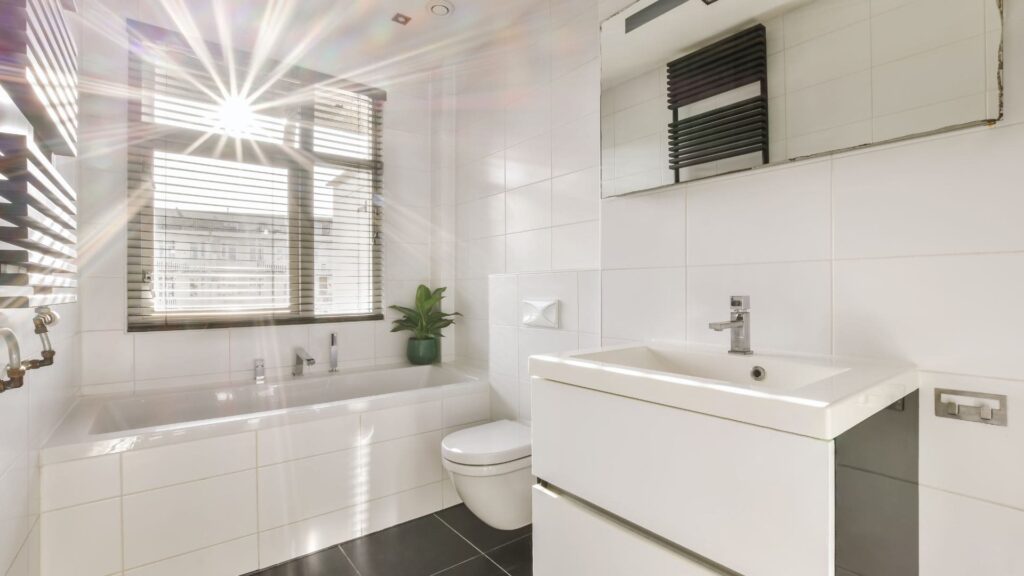 Baño moderno y minimalista con bañera, lavabo con espejo grande, inodoro y planta decorativa en un apartamento vacacional en la Costa del Sol. La luz natural entra por la ventana con persianas, iluminando la estancia.